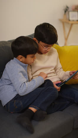 Vertical-Video-Of-Two-Young-Boys-Sitting-On-Sofa-At-Home-Playing-Games-Or-Streaming-Onto-Digital-Tablet-Together-2